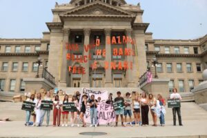 Youth Salmon Protectors stand with a banner in front of a dam built from cardboard boxes on the Idaho State Capitol steps.